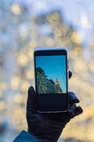 mano della donna in un guanto di pelle nera che tiene uno smartphone nero per scattare foto della città di Natale in una soleggiata giornata invernale. sfondo bokeh