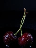 Due maturo ciliegia frutti di bosco con gocce di acqua su il sbucciare su un' nero sfondo. foto