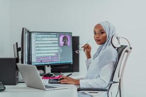 giovane afro americano moderno musulmano donna d'affari indossare un' sciarpa nel un' creativo luminosa ufficio posto di lavoro con un' grande schermo. foto