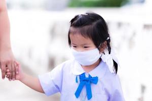 asiatico prescolastico bambino ragazza indossare un' protettivo viso maschera. ragazzo indossare scuola uniforme a piedi e lei hold mano nel mano madre. dolce sorridente sotto il bianca maschera. indietro per scuola concetto. foto