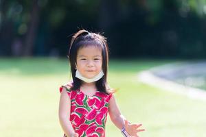 carino asiatico ragazza dolce Sorridi. bambino prende viso maschera sotto il suo mento. luminosa verde prato sfondo. estate o primavera. contento ragazzo vestito nel rosso guardato a telecamera, nel caldo luce del sole. bambini 3-4 anni vecchio. foto