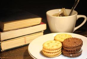 tazza di tè con piccolo cucchiaio, biscotto su un' piatto e un' pila di libri su il tavolo selettivo messa a fuoco foto