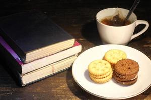 tazza di tè con piccolo cucchiaio, biscotto su un' piatto e un' pila di libri su il tavolo selettivo messa a fuoco foto