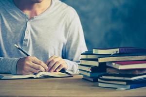 vicino su di un' uomo scrittura nel taccuino, pila di vecchio libri su di legno scrivania. Vintage ▾ tono foto