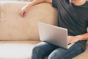 vicino su di casuale giovane uomo seduta su divano e utilizzando un' il computer portatile a casa. foto