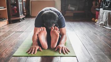 giovane asiatico uomo praticante yoga, seduta nel seduto inoltrare piegare esercizio, paschimottanasana posa su verde stuoia yoga a di legno casa. salutare vivente foto