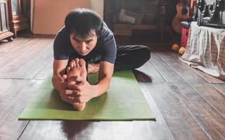 giovane asiatico uomo praticante yoga nel un' di legno camera seduta nel posa janu sirsasana su un' verde yoga stuoia a un' di legno Casa. salutare vita concetto foto