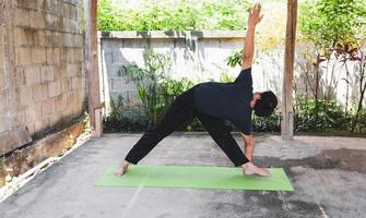 salutare vivente concetto di giovane asiatico uomo praticante yoga asana triangolo posa, Lavorando fuori, pose su un' verde yoga stuoia. al di fuori esercizio nel il giardino. salutare stile di vita. foto