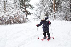 i piedi dei bambini con gli sci di plastica rossa con i bastoncini attraversano la neve da uno scivolo: uno sport invernale, intrattenimento per famiglie all'aria aperta. una bambina scivola giù per il pendio fin dalla tenera età. copia spazio foto