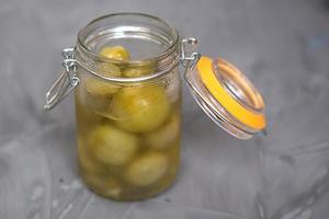 sottaceto verde piccolo pomodori nel un' bicchiere vaso con un' coperchio su un' grigio sfondo. salato ciliegie, fatti in casa, olive nel salamoia. foto