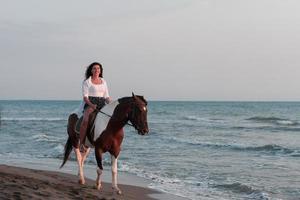 donna nel estate Abiti gode equitazione un' cavallo su un' bellissimo sabbioso spiaggia a tramonto. selettivo messa a fuoco foto