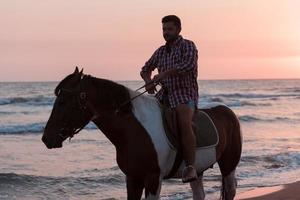 un' moderno uomo nel estate Abiti gode equitazione un' cavallo su un' bellissimo sabbioso spiaggia a tramonto. selettivo messa a fuoco foto