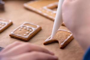 la giovane donna sta decorando i biscotti della casa di pan di zenzero di natale biscotto a casa con guarnizione di glassa in un sacchetto di glassa, primo piano, stile di vita. foto