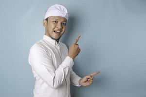 eccitato balinese uomo indossare udeng o tradizionale fascia per capelli e bianca camicia puntamento a il copia spazio accanto lui, isolato di blu sfondo foto