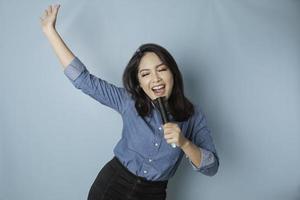ritratto di spensierato asiatico donna, avendo divertimento karaoke, cantando nel microfono mentre in piedi al di sopra di blu sfondo foto