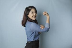 eccitato asiatico donna indossare un' blu camicia mostrando forte gesto di sollevamento sua braccia e muscoli sorridente con orgoglio foto