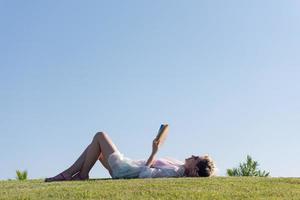 donna dire bugie e lettura sua preferito libro su un' prato coperto con fresco verde erba su un' soleggiato estate o primavera giorno. foto
