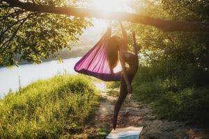 un' giovane femmina ginnasta è impegnato nel aereo yoga, utilizzando un' combinazione di tradizionale yoga pose, pilates e danza utilizzando un' amaca a tramonto nel natura. salutare stile di vita. foto