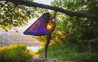 un' giovane femmina ginnasta è impegnato nel aereo yoga, utilizzando un' combinazione di tradizionale yoga pose, pilates e danza utilizzando un' amaca a tramonto nel natura. salutare stile di vita. foto