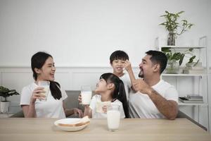 un' salutare asiatico tailandese famiglia, poco figli, e giovane genitori bevanda fresco bianca latte nel bicchiere e pane gioia insieme a un' cenare tavolo nel mattina, benessere nutrizione casa prima colazione pasto stile di vita. foto