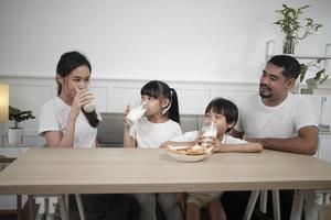 un' salutare asiatico tailandese famiglia, poco figli, e giovane genitori bevanda fresco bianca latte nel bicchiere e pane gioia insieme a un' cenare tavolo nel mattina, benessere nutrizione casa prima colazione pasto stile di vita. foto