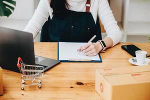 imprenditore proprietario utilizzando penna punto Nota e il computer portatile controllo in linea Acquista shopping ordine per preparazione imballare Prodotto scatola. Imballaggio merce per consegna per cliente. foto