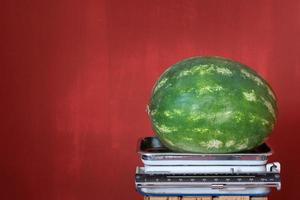 un vecchio cucina scala sta contro un' rosso sfondo con spazio per testo. Là è un' grande verde anguria su il bilancia foto