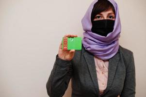 ritratto di giovane musulmano donna indossare formale indossare, proteggere viso maschera e hijab testa sciarpa, hold turkmenistan bandiera carta contro isolato sfondo. coronavirus nazione concetto. foto