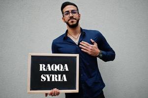 arabo uomo indossare blu camicia e occhiali hold tavola con raqqa Siria iscrizione. maggiore città nel islamico mondo concetto. foto