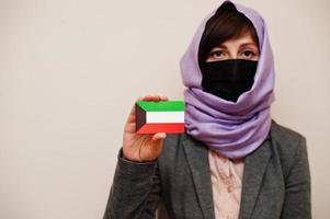 ritratto di giovane musulmano donna indossare formale indossare, proteggere viso maschera e hijab testa sciarpa, hold Kuwait bandiera carta contro isolato sfondo. coronavirus nazione concetto. foto