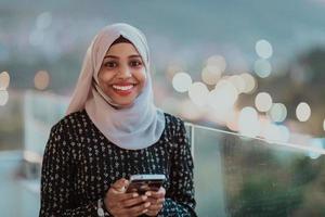 giovane musulmano donna indossare sciarpa velo su urbano città strada a notte sms su un' smartphone con bokeh città leggero nel il sfondo. foto