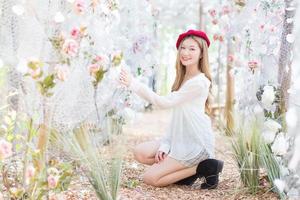 la donna asiatica ha lunghi capelli color bronzo e indossa un berretto rosso, un vestito bianco e stivali neri mentre si siede in un giardino fiorito nella stagione invernale. foto