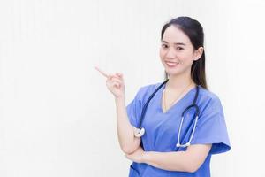 asiatico bellissimo donna medico nel un' blu uniforme sta e sorrisi mentre puntamento per il superiore su un' bianca sfondo. foto