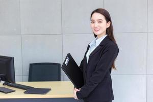 asiatico professionale Lavorando femmina nel un' nero completo da uomo detiene appunti nel sua mani e fiducioso sorrisi nel ufficio camera. foto