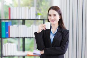 asiatico attività commerciale donna chi indossa blu camicia e nero completo da uomo detiene caffè tazza nel sua mano e Sorridi felicemente a opera ufficio come sfondo. foto