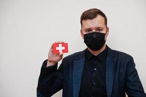 europeo uomo indossare nero formale e proteggere viso maschera, hold Svizzera bandiera carta isolato su bianca sfondo. Europa coronavirus covid nazione concetto. foto