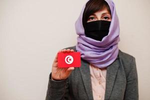 ritratto di giovane musulmano donna indossare formale indossare, proteggere viso maschera e hijab testa sciarpa, hold tunisia bandiera carta contro isolato sfondo. coronavirus nazione concetto. foto