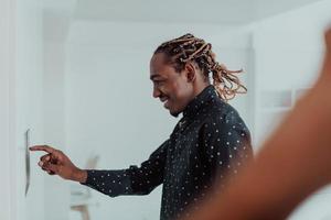 sorridente africano americano uomo utilizzando moderno inteligente casa sistema, controllore su parete, positivo giovane uomo commutazione temperatura su termostato o attivazione sicurezza allarme nel appartamento foto