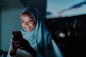 giovane musulmano donna indossare sciarpa velo su urbano città strada a notte sms su un' smartphone con bokeh città leggero nel il sfondo.