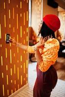 elegante afro americano donna nel rosso francese berretto, grande oro collo catena polka punto camicetta e pelle pantaloni conduce un' vivere trasmissione di mobile Telefono per sociale Rete. foto