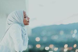 africano musulmano donna nel il notte su un' balcone sorridente a il telecamera con città bokeh luci nel il sfondo. foto