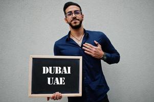 arabo uomo indossare blu camicia e occhiali hold tavola con dubai Emirati Arabi Uniti iscrizione. maggiore città nel islamico mondo concetto. foto