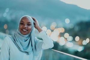 africano musulmano donna nel il notte su un' balcone sorridente a il telecamera con città bokeh luci nel il sfondo. foto