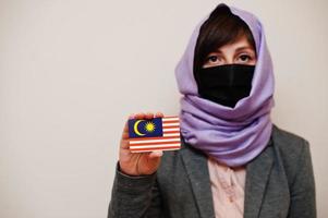 ritratto di giovane musulmano donna indossare formale indossare, proteggere viso maschera e hijab testa sciarpa, hold Malaysia bandiera carta contro isolato sfondo. coronavirus nazione concetto. foto