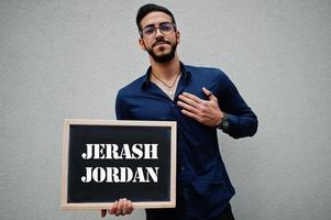 arabo uomo indossare blu camicia e occhiali hold tavola con jerash Giordania iscrizione. maggiore città nel islamico mondo concetto. foto