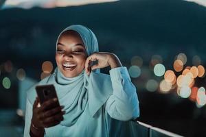 giovane musulmano donna indossare sciarpa velo su urbano città strada a notte sms su un' smartphone con bokeh città leggero nel il sfondo. foto