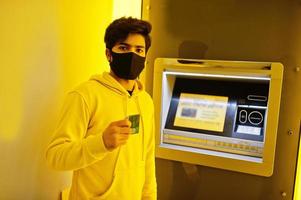 urbano giovane fricchettone indiano uomo nel un' alla moda giallo felpa a banca. freddo Sud asiatico tipo indossare felpa con cappuccio e nero viso proteggere maschera, durante nuovo normale, si ritira denaro contante a ATM. foto