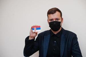 europeo uomo indossare nero formale e proteggere viso maschera, hold Serbia bandiera carta isolato su bianca sfondo. Europa coronavirus covid nazione concetto. foto