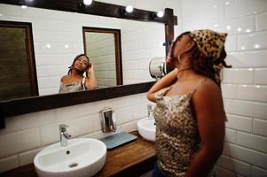 bello africano americano donna con dreadlocks nel leopardo attrezzatura guardare nel il specchio a bagno bar. bellissimo freddo alla moda nero giovane ragazza interno. foto