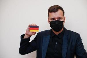 europeo uomo indossare nero formale e proteggere viso maschera, hold Germania bandiera carta isolato su bianca sfondo. Europa coronavirus covid nazione concetto. foto